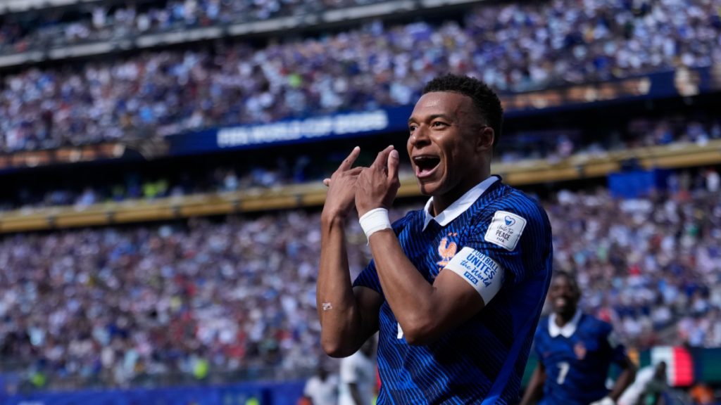 Kylian Mbappé comemora um gol pela seleção da França durante uma partida, sorrindo e levantando as mãos no estádio com a torcida ao fundo, tema jogos da Copa hoje.