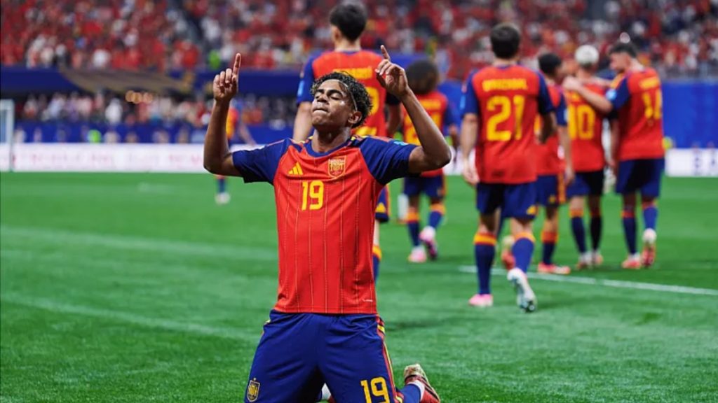 Yamal comemora em campo com a camisa da Espanha, braços levantados, enquanto o placar ao fundo mostra partida de futebol e outros jogadores ao redor, em comparação com craque Messi.