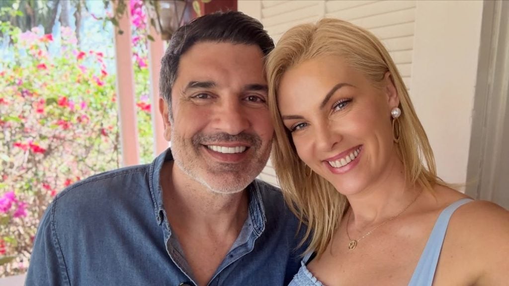 Ana Hickmann ao lado de Edu Guedes em foto sorridente, com os dois abraçados em ambiente interno, iluminação natural e decoração ao fundo com flores.