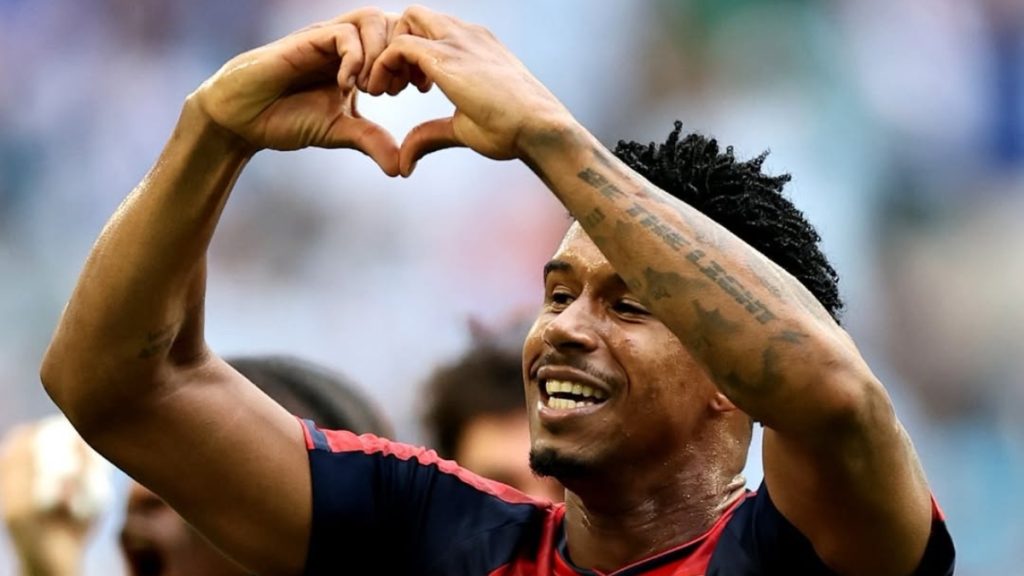 Jogador de futebol com camiseta escura levanta os braços formando um coração com as mãos durante uma partida no estádio. Cabo Verde não aparece na imagem.