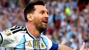 Messi em partida de futebol, em primeiro plano, comemorando com a boca aberta e o olhar para o lado, usando uniforme da Argentina, em estÃ¡dio cheio.