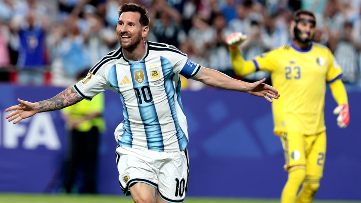 Lionel Messi em campo com a camisa da Argentina, comemorando com os braços abertos durante uma partida em estádio lotado, enquanto um jogador do time adversário aparece desfocado ao fundo. Messi faz história e se isola como maior artilheiro das Copas.