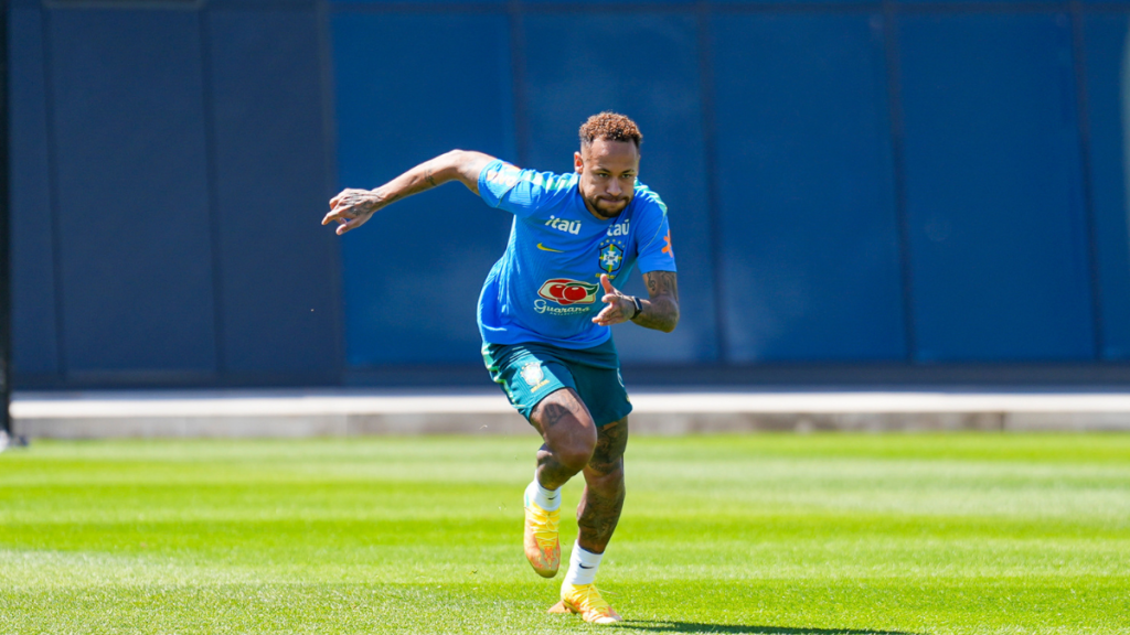 Neymar em treino da Seleção Brasileira, com camisa azul e escudo do Brasil, correndo em campo com expressão concentrada durante atividade física ao ar livre.