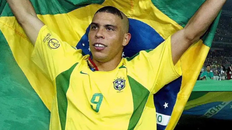 Camisa do Brasil de 2002 em destaque: jogador em uniforme amarelo com escudo da seleção, número 9 e bandeira do Brasil ao fundo, em cena comemorativa.