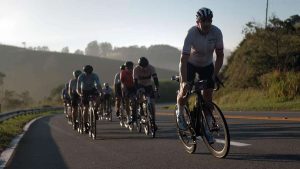 Ciclistas do Tour de France pedalam em uma estrada no interior, com corredor da prova em primeiro plano e pelotão ao fundo, em rota que passa por Serra Negra no Circuito das Águas Paulista.