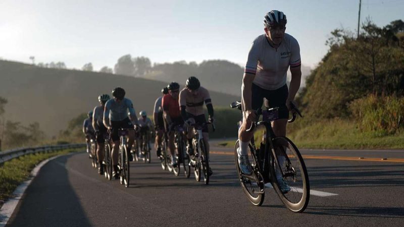 Ciclistas do Tour de France pedalam em uma estrada no interior, com corredor da prova em primeiro plano e pelotão ao fundo, em rota que passa por Serra Negra no Circuito das Águas Paulista.