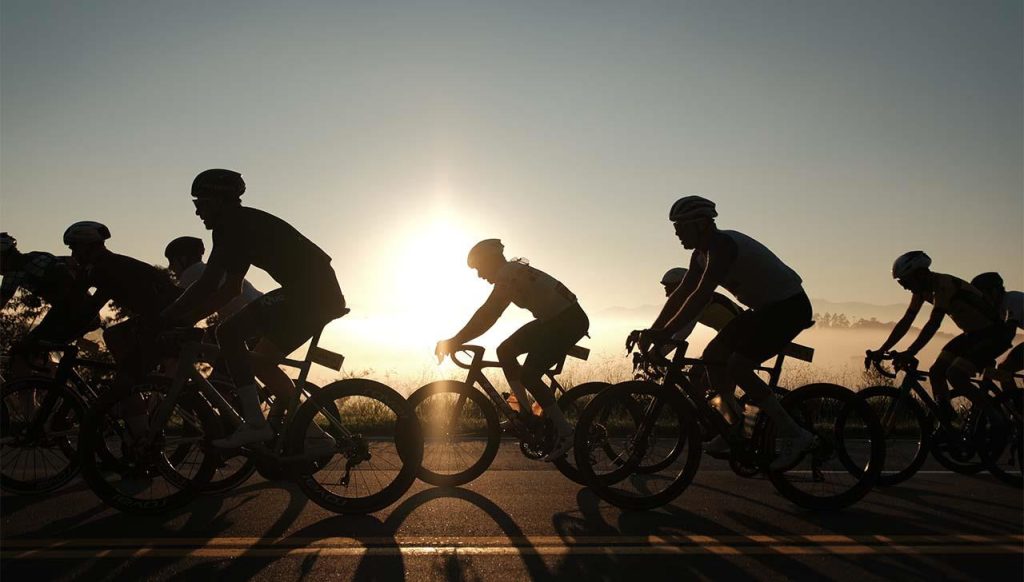 Ciclistas em fila no fim de tarde durante a Serra Negra Tour de France, com o sol baixo ao fundo e percursos indicados de 54 km e 99 km