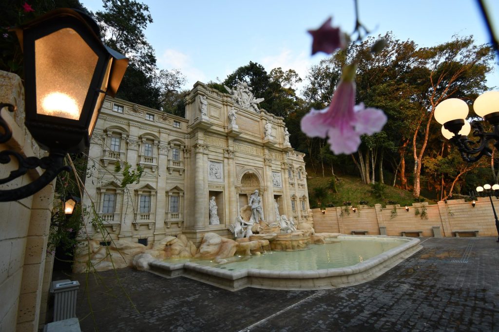 Luz externa e jardim com fonte ao lado de um prédio histórico e arcos, em clima de evento, com árvores e bandeiras ao fundo, remetendo à Serra Negra Tour de France.