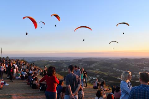 Turistas e participantes assistem a uma etapa com paraquedas no pôr do sol em Serra Negra, destino do Serra Negra Tour de France, com céu e montanhas ao fundo.