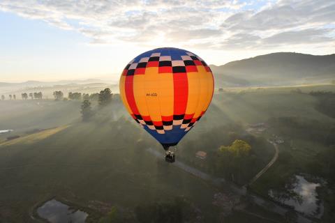 Balão de ar quente em voo sobre paisagens rurais e montanhosas em Serra Negra, com clima de visibilidade internacional ligado ao Tour de France e à competição.