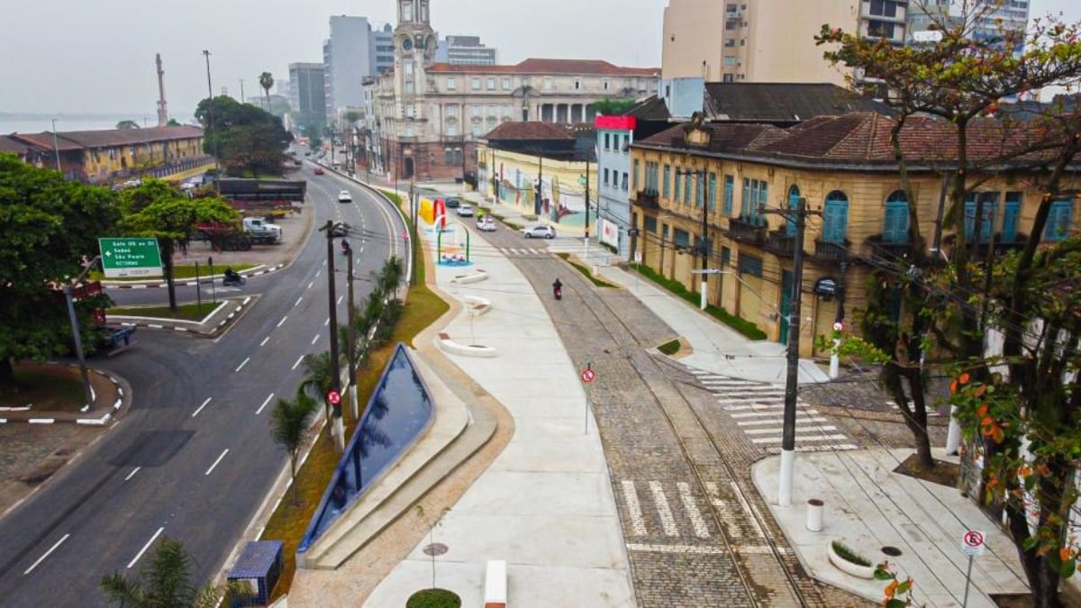 Vista de uma avenida em Santos com vias urbanas, calçadas e faixa de pedestres. No cenário, destaca-se a possibilidade de ativar Zona Azul por QR Code em comércios, orientada aos motoristas.