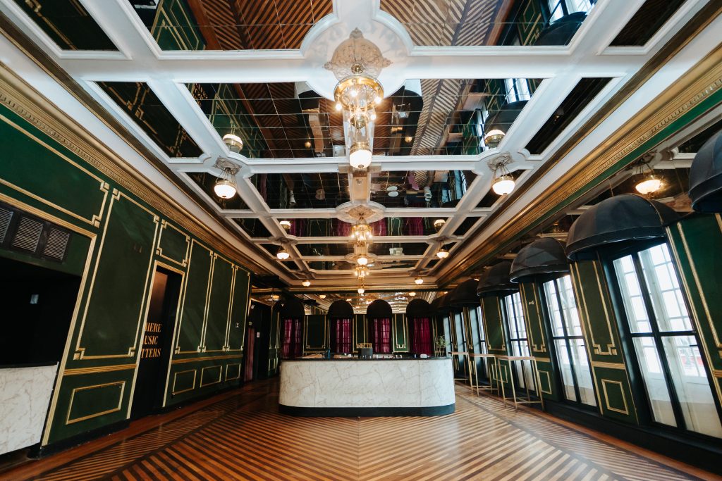 Interior do Jockey Club Campineiro com corredor elegante, piso de madeira e mármore, iluminação ornamentada e vitrais nas laterais, dando destaque ao ambiente histórico.