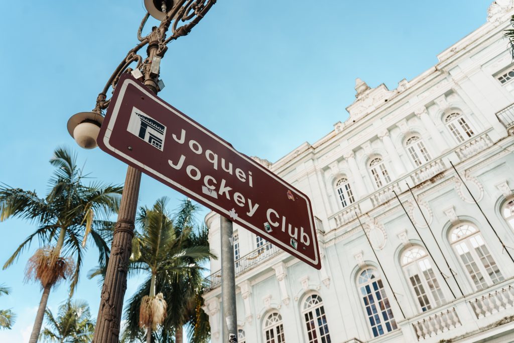 Placa de sinalização do Jockey Club Campineiro, com o texto “Jockey Club” e edifício histórico branco ao fundo, sob céu azul e palmeiras ao lado.
