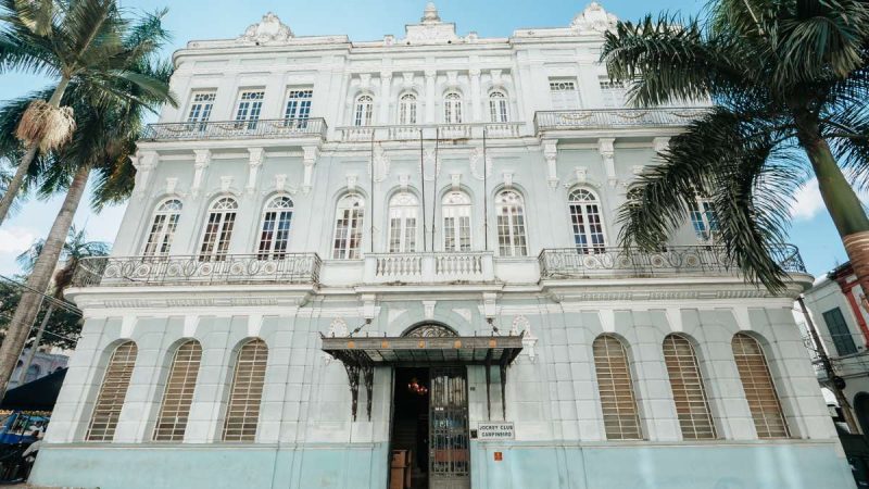 Fachada do Jockey Club Campineiro, com prédio histórico branco, palmeiras ao redor e entrada principal ao centro em foto para livro e exposição