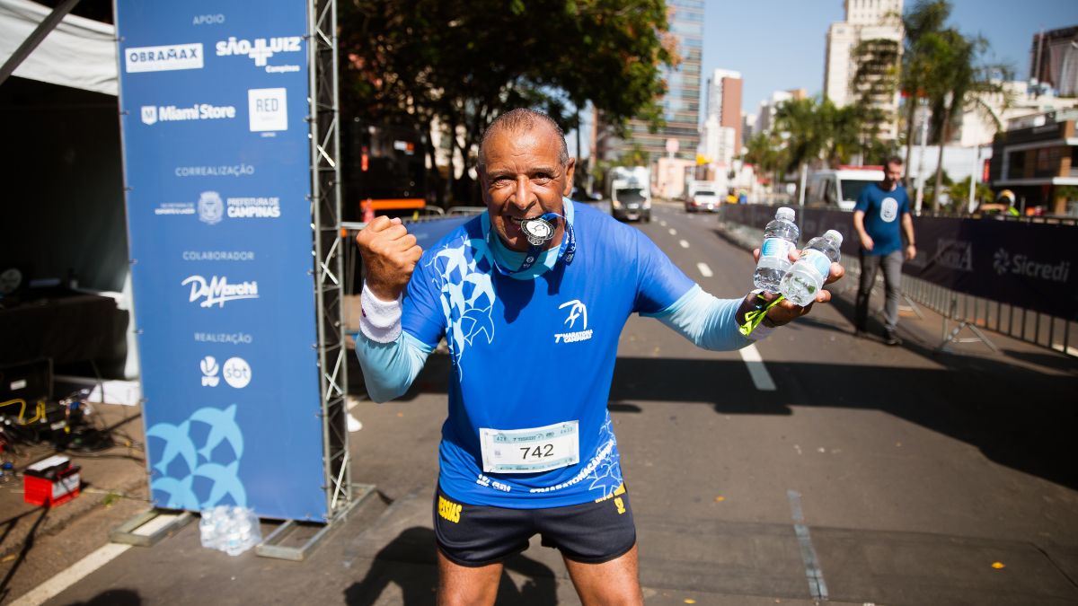 Corredor em prova na cidade, com medalha e garrafas de água na linha de chegada, usando número 742 e camiseta azul, em um cenário urbano que remete à Maratona de Campinas. Último dia de inscrições hoje.