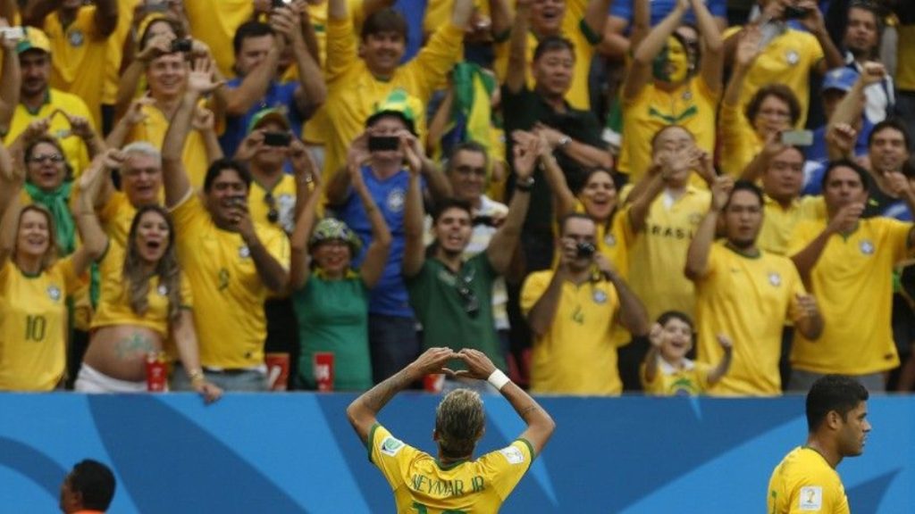 Neymar em destaque durante partida de futebol, com a camisa da seleção brasileira amarela, enquanto o público ao fundo vibra e torcedores levantam as mãos.