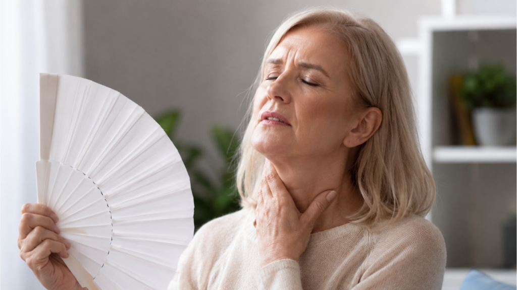 Mulher na fase da menopausa segura um leque aberto e apresenta expressão de desconforto, com a mão no pescoço. Cena interna que ilustra sintomas controlados por nova medicação não hormonal.