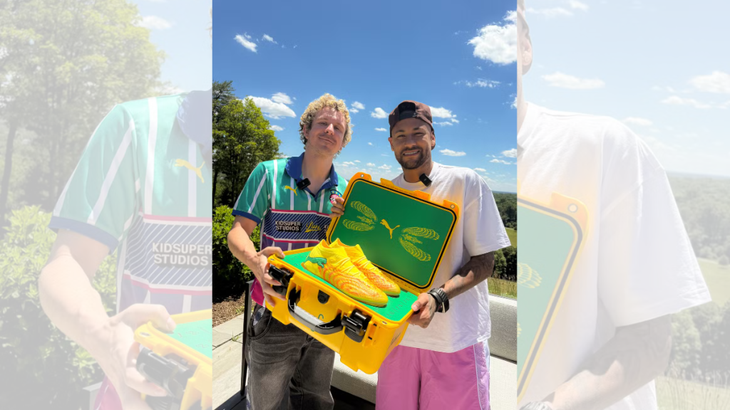 Entrega da nova chuteira da Seleção para Neymar: jogador de cabelo claro segura chuteiras amarelas e caixa verde e amarela, enquanto uma segunda pessoa acompanha ao ar livre sob céu azul.