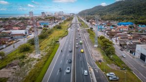 Vista aérea de uma rodovia com faixas e veículos em circulação, passando por área urbana e vegetação ao lado do litoral e de colinas, ilustrando investimento nas rodovias da Baixada Santista e Vale do Ribeira com R$ 260 milhões