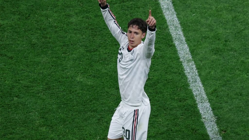 Jogador da seleção mexicana em campo, vestindo uniforme branco e acenando com o braço na comemoração, em partida que garantiu campanha perfeita no Grupo A e homenagem a Guillermo Ochoa.