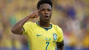 Vinícius Júnior em destaque com a camisa amarela da Seleção Brasileira, fazendo gesto de comemoração em partida da Copa, liderando a nova fase do time.