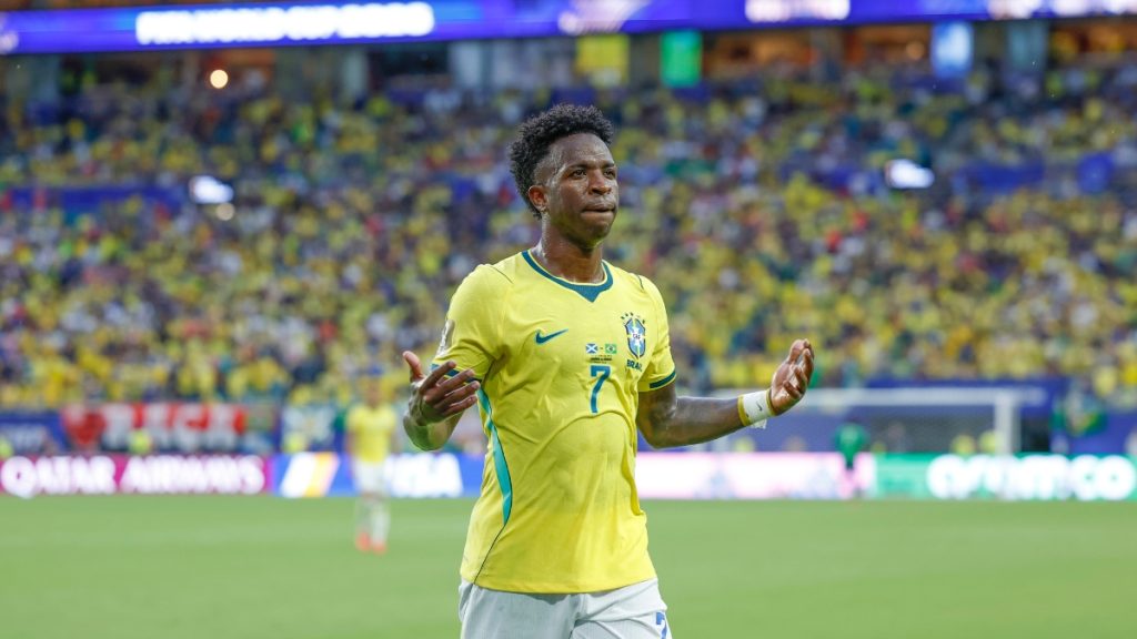 Vinícius Júnior em campo com a camisa da seleção brasileira amarela durante uma partida, em meio a um estádio lotado e torcida ao fundo