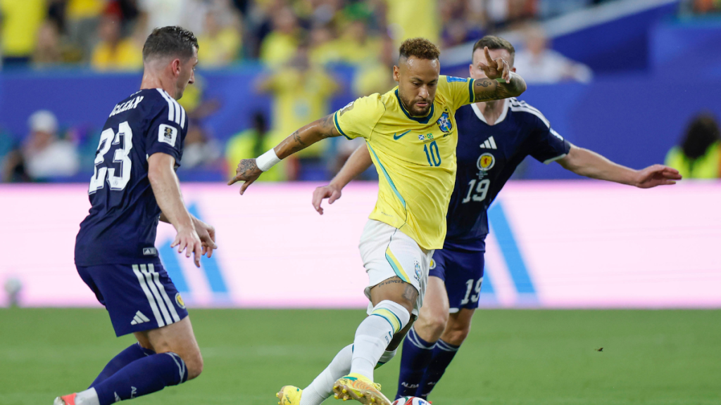 Neymar em ação na partida do Brasil contra a Escócia, driblando e tentando avançar com a bola durante o jogo, com jogadores adversários ao redor.