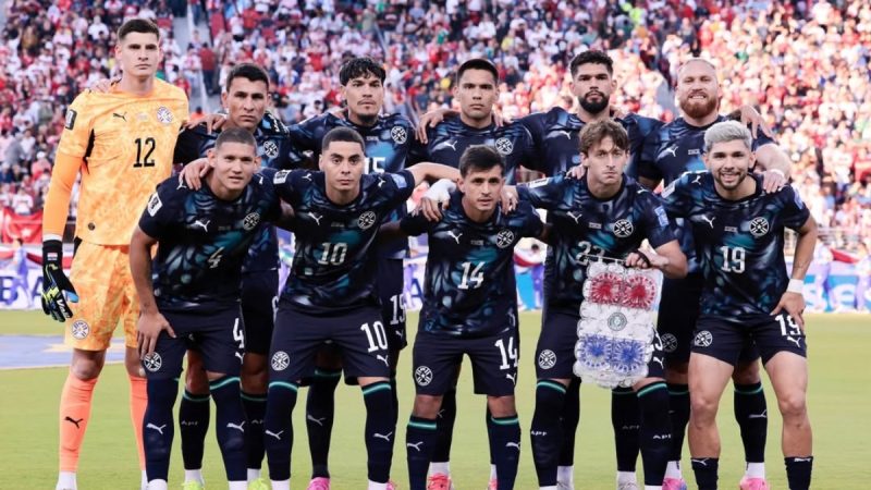 Elenco da seleção do Paraguai em campo durante partida decisiva da Copa do Mundo, com jogadores em uniforme escuro e goleiro usando uniforme laranja, diante de torcida lotada.