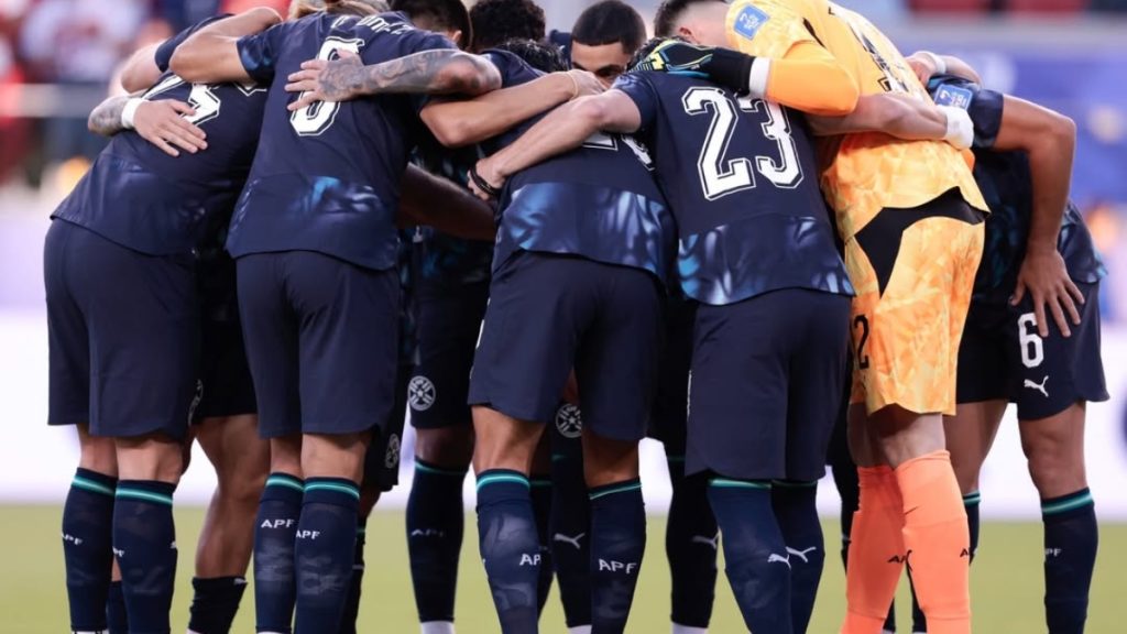 Paraguai aposta em base com ligações ao Brasil: jogadores se reúnem em abraço no campo durante treino ou partida, formando um grupo de comemoração em uniforme azul e amarelo com número 23.