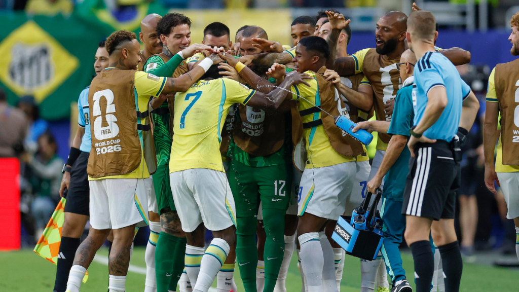 Neymar da seleção brasileira comemora com Vini Jr. e o restante do time após um gol contra a Escócia, em clima de euforia e abraço coletivo.