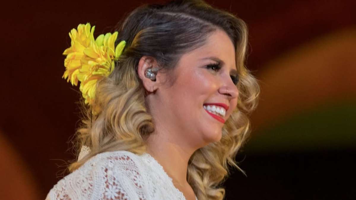 Imagem em close de uma mulher sorridente com cabelo loiro ondulado, segurando ou usando um arranjo amarelo na orelha e vestindo roupa clara, em destaque contra fundo escuro; referência ao filme sobre Marília Mendonça anunciando o tema da produção
