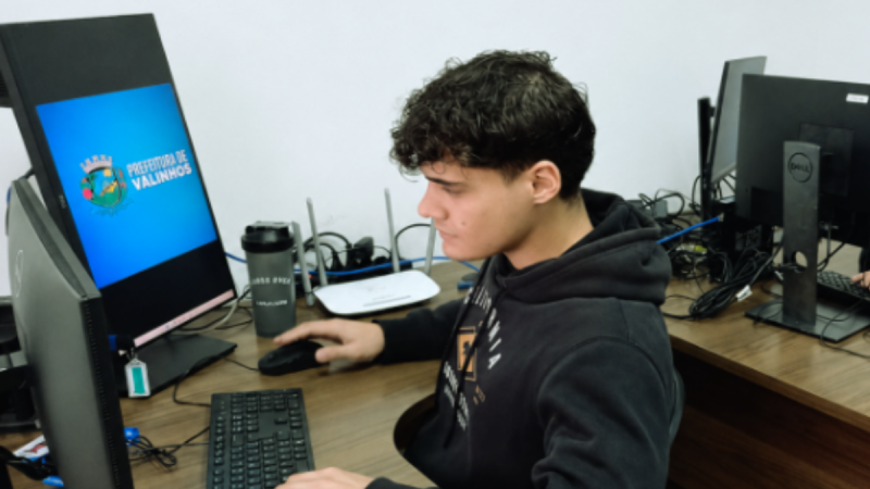 Jovem estudante trabalhando em computador em um escritório durante seleção de vagas de estágio em Valinhos. Tela do monitor com identidade da Prefeitura de Valinhos ao fundo.