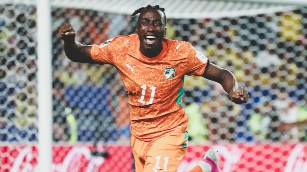 Jogador de futebol em campo, usando uniforme laranja com o número 11 e escudo verde, comemorando com os braços abertos durante uma partida em um estádio