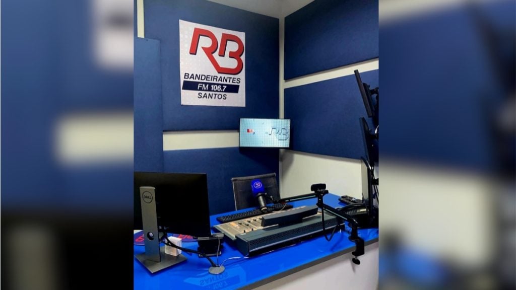 Estúdio de rádio com mesa de som, microfones e painel azul, com o logo da Rádio Bandeirantes FM 106.7 em Santos. Rádio Bandeirantes Baixada Santista