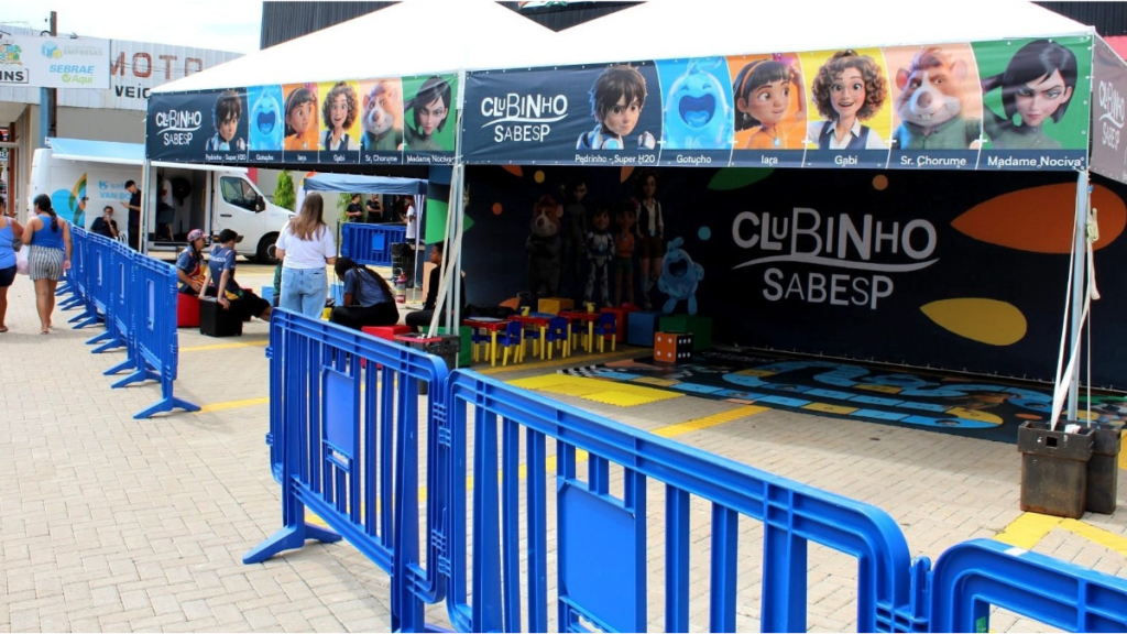 Caravana Sabesp Guarujá com o Clubinho Sabesp, tenda colorida e área de jogos para crianças em evento externo, com cadeiras e atividades educativas sobre uso consciente da água.
