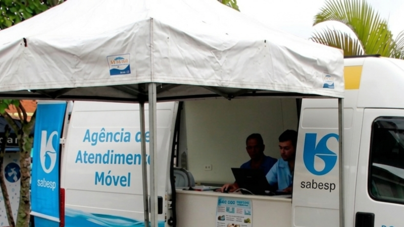 Caravana Sabesp Guarujá com tenda de Agência de Atendimento Móvel e van da Sabesp, onde equipes atendem ao lado de computadores durante ação no bairro.