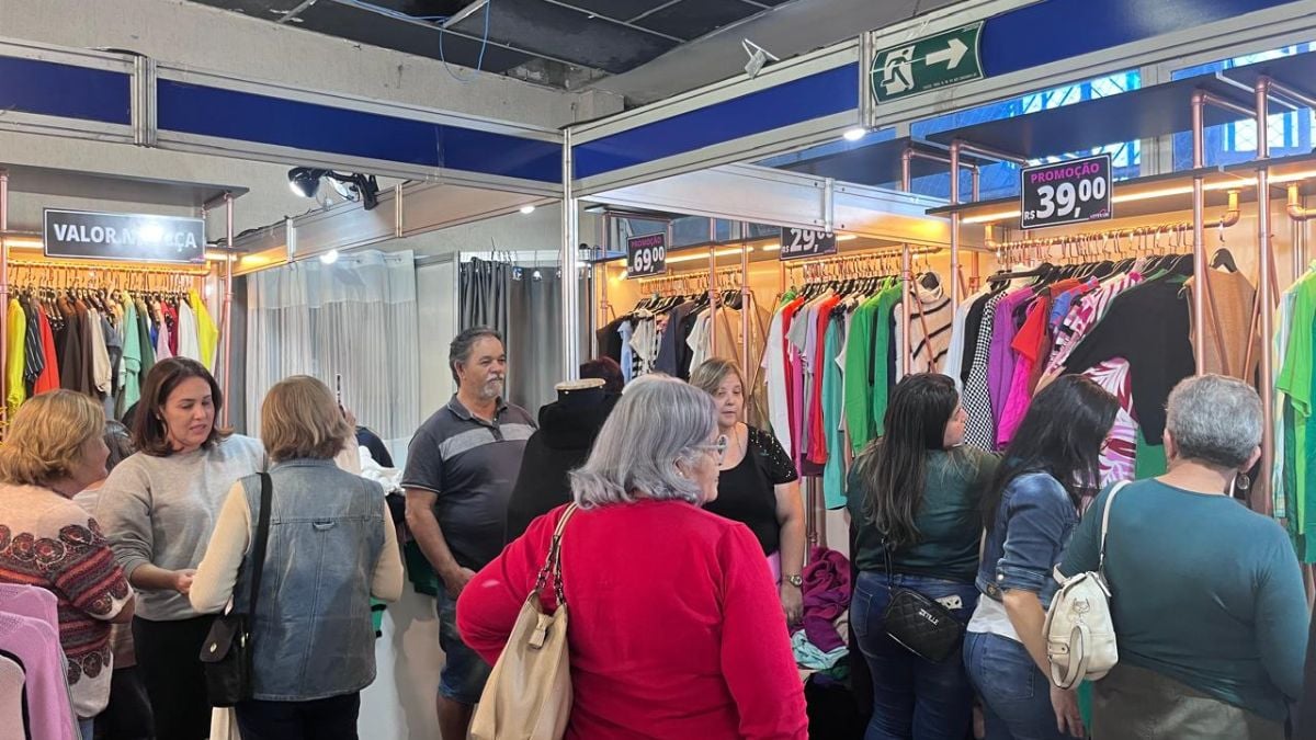 Feimalhas em Santos apresenta novidades de inverno em feira de moda: pessoas visitam um estande com araras de roupas coloridas e faixas de preços de promoções no ambiente coberto.