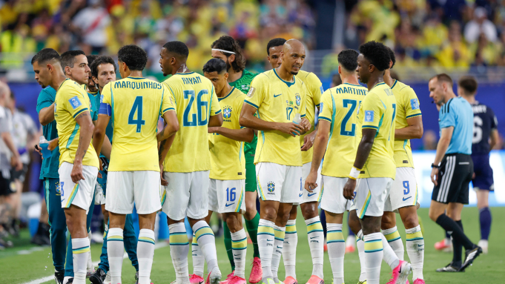 Seleção brasileira em campo reunida em atitude de concentração durante partida da Copa, com jogadores com a camisa amarela e escudos do Brasil, prestes a enfrentar o Japão na próxima fase.
