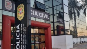 Prédio da Polícia Civil com placa “POLICIA CIVIL” ao fundo e entrada com estrutura vermelha, em Americana; imagem relacionada à prisão de casal por suspeita de maus-tratos contra filho autista
