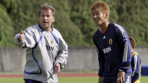 Treino de futebol ao ar livre com dois técnicos orientando atletas: um treinador de agasalho branco e outro em uniforme azul, em cenário que ilustra o Plano de 100 Anos da JFA para o Japão