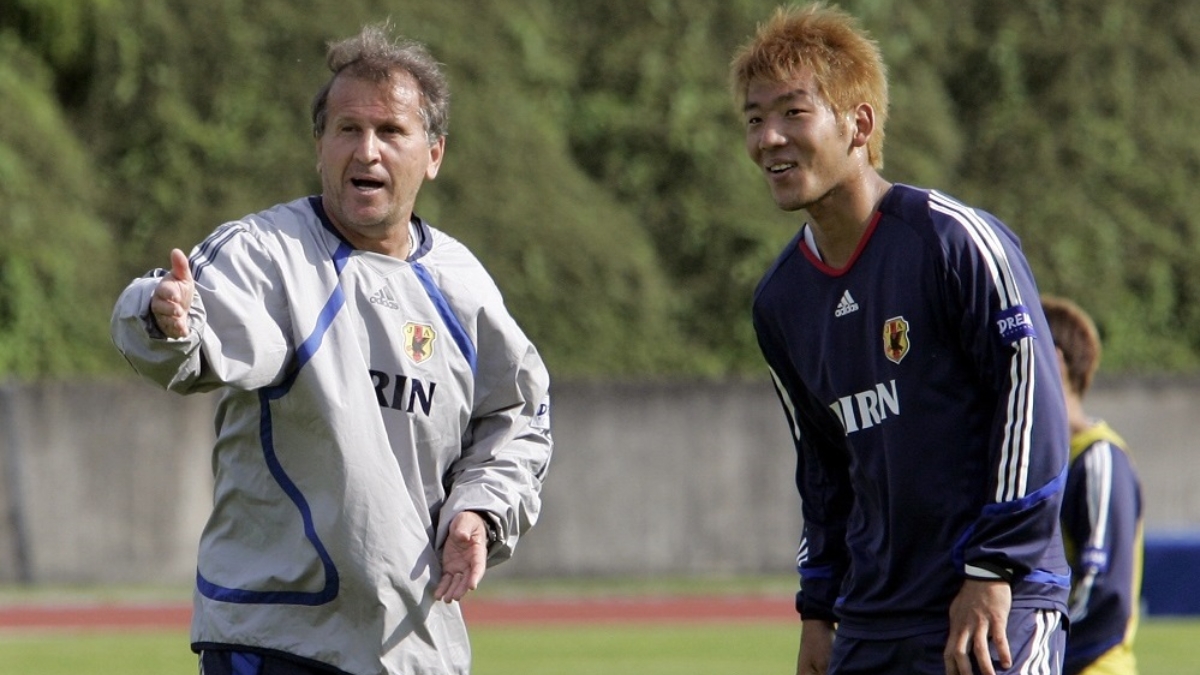 Treino de futebol ao ar livre com dois técnicos orientando atletas: um treinador de agasalho branco e outro em uniforme azul, em cenário que ilustra o Plano de 100 Anos da JFA para o Japão
