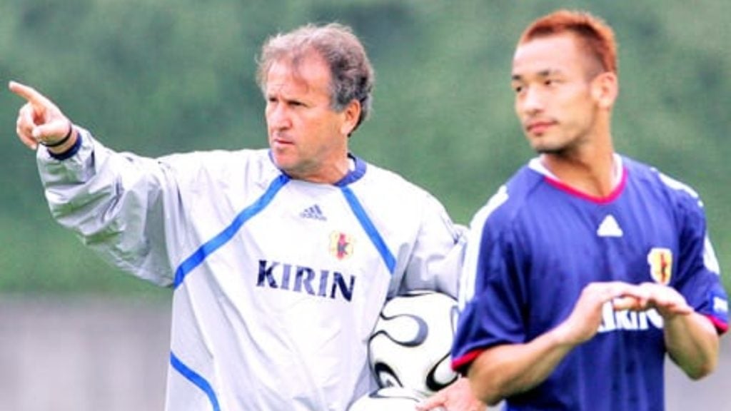 Treinador japonês orienta jogadores durante treino em campo, com uniforme branco escrito “KIRIN” e atleta de azul, ilustrando o Plano de 100 Anos da JFA para dominar o futebol no Japão.