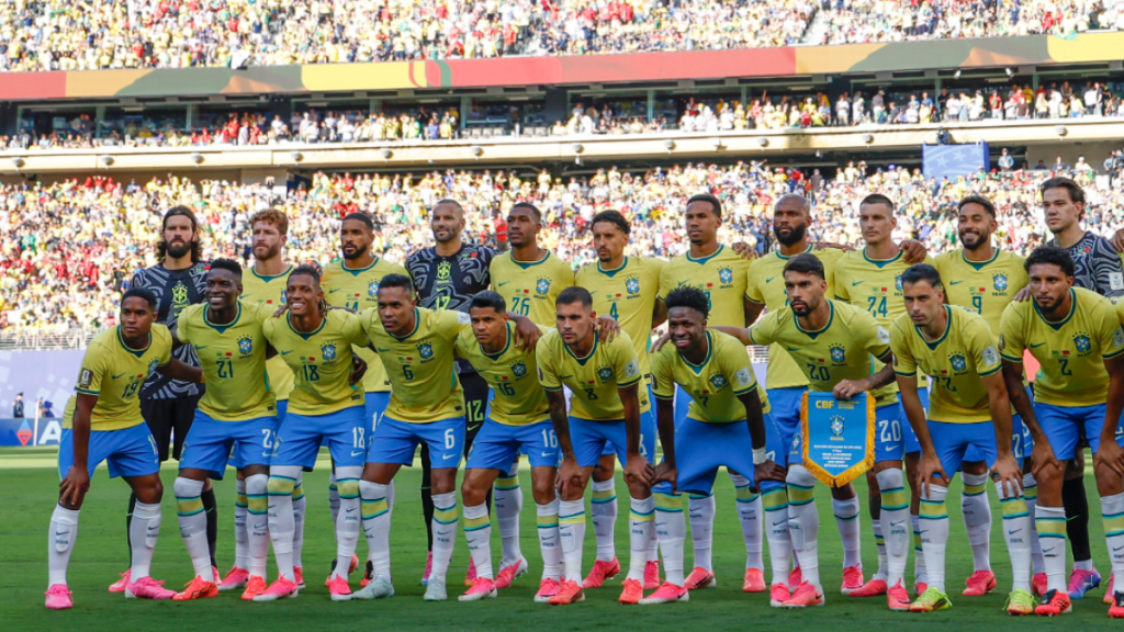 Seleção Brasileira em foto coletiva, uniforme amarelo e azul, em campo com torcida ao fundo, em preparação para o confronto Brasil x Japão pela Copa do Mundo 2026.