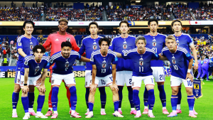 Seleção do Japão em campo com uniforme azul e branco, formando um time antes da partida; torcedores aparecem ao fundo, destacando a tensão da Copa entre Japão e Brasil.
