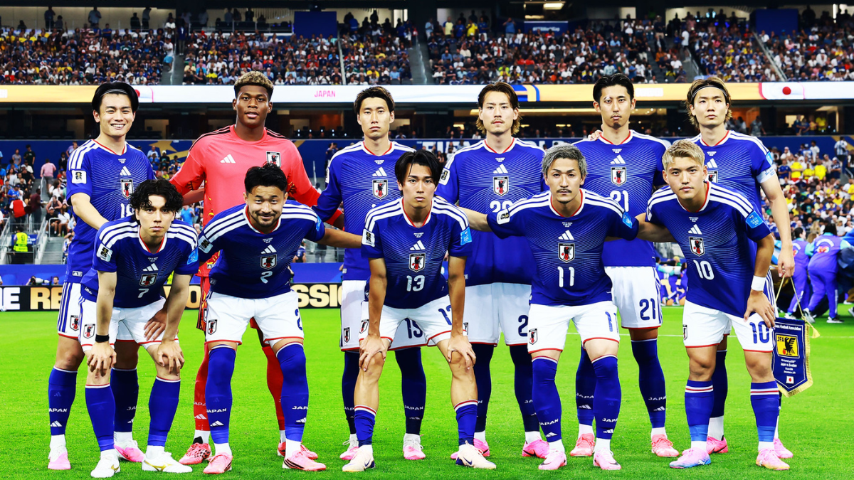 Seleção do Japão em campo com uniforme azul e branco, formando um time antes da partida; torcedores aparecem ao fundo, destacando a tensão da Copa entre Japão e Brasil.