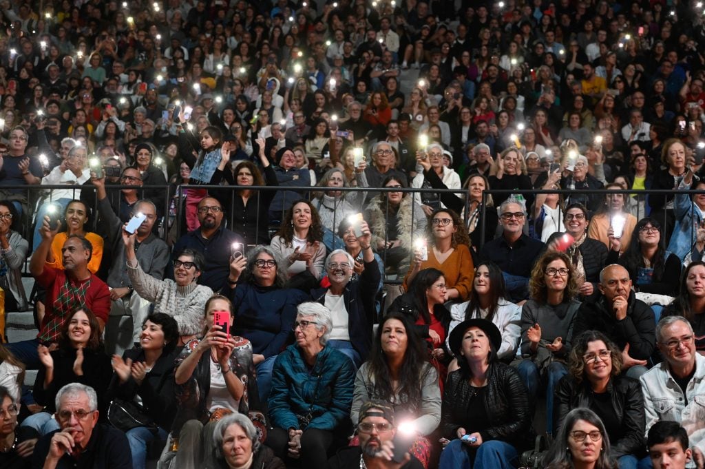 Público em um teatro lotado aplaudindo e levantando celulares com luzes durante um evento musical, com muitas pessoas sentadas em fileiras e flashes de fotografias ao fundo.