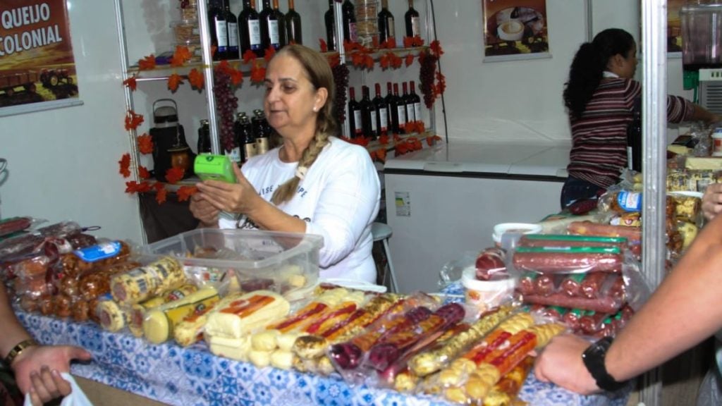 Feimalhas em Santos: banca de gastronomia de inverno com variedade de doces e enfeites, onde uma atendente prepara e organiza os produtos em embalagens e bandejas ao lado de bebidas.