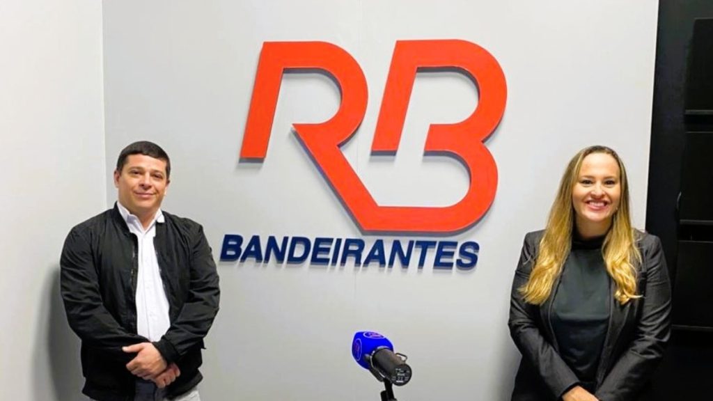 Rádio Bandeirantes Baixada Santista: Rafaella Ferreira e Osvaldinho Stuchi posam em estúdio com o logo da emissora ao fundo.