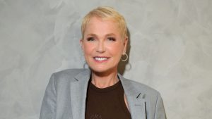 Xuxa em foto de estúdio, sorridente, com cabelo curto loiro e blazer cinza, em clima de ensaio para a turnê de despedida com as Paquitas.
