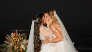 Lauana Prado e Tati Dias trocam um beijo no dia do casamento, com véu e vestido branco, em cerimônia ao ar livre com vista para o mar ao fundo.